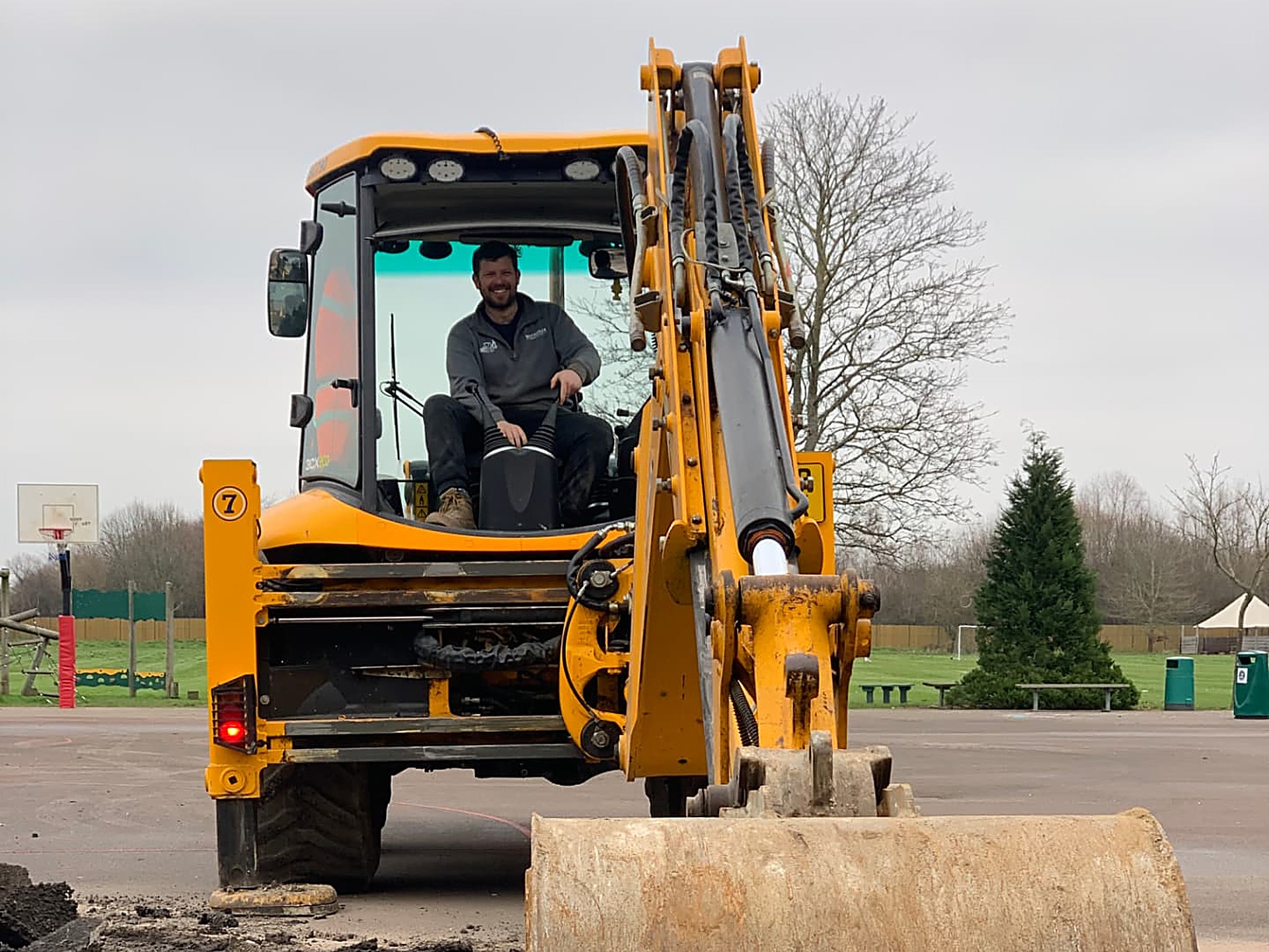 A yellow backhoe loader with its bucket in a trench on a paved recreation ground, with a basketball hoop and trees behind