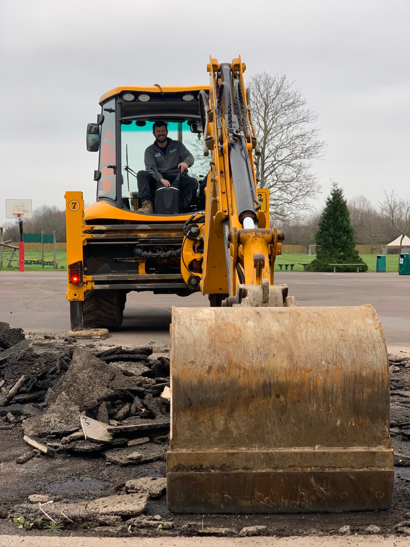 A yellow backhoe loader with its bucket lowered beside broken asphalt on a paved playground, with a basketball hoop and trees behind